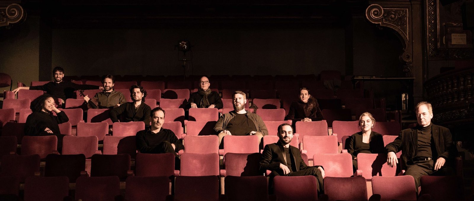 Twelve members of the Europa Meta Orchestra sitting among the red velvet seats of a theatre, in warm, contrasting light