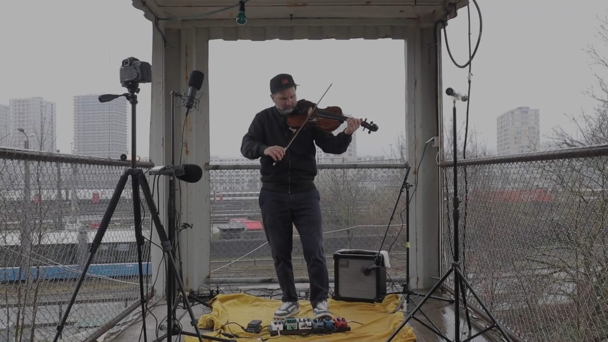 Diego Kohn playing viola in an open shelter above a railway yard in Zürich, surrounded by microphones and pedals