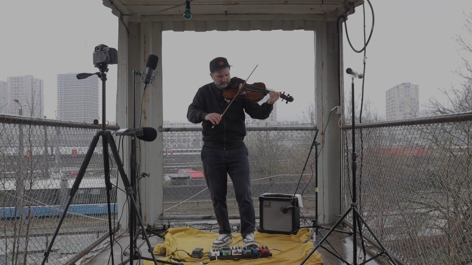 Diego Kohn playing viola in an open shelter above a railway yard in Zürich, surrounded by microphones, a small amplifier and effect pedals