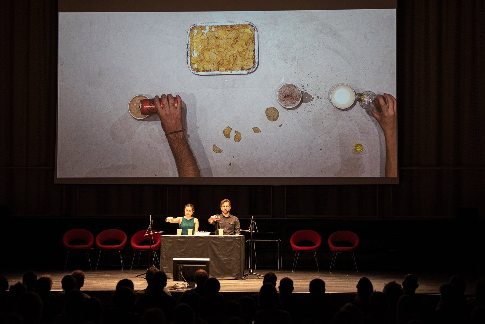 Gemma Galeano and Diego Kohn at a table on stage, pouring drinks; above them a large screen shows their hands from above among cans, cups and a tray of crisps