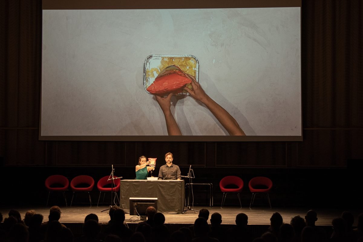 On the screen, hands pour crisps from a red bag into an aluminium tray; the two performers sit at the table below