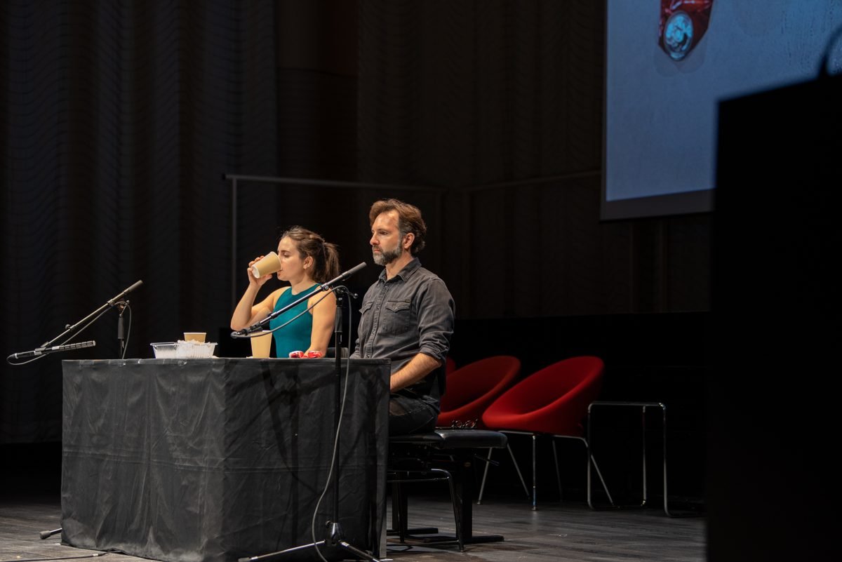 Gemma Galeano drinking from a paper cup next to Diego Kohn at the table, microphones in front of them