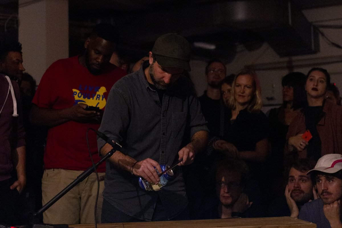 Diego Kohn opening a can at a wooden board with a microphone, audience close around him