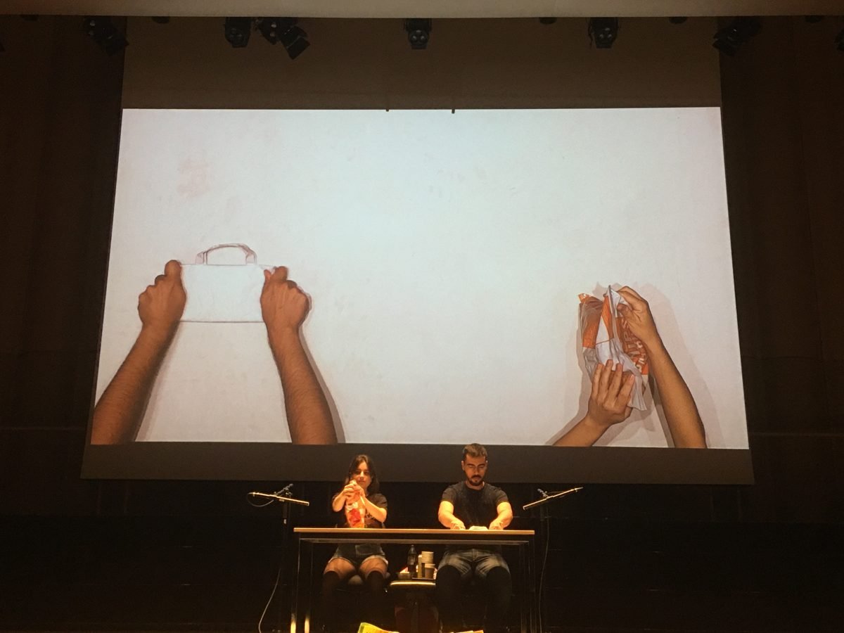 Rehearsal: on the screen, hands hold an empty tray and a plastic wrapper; the performers sit at the table below