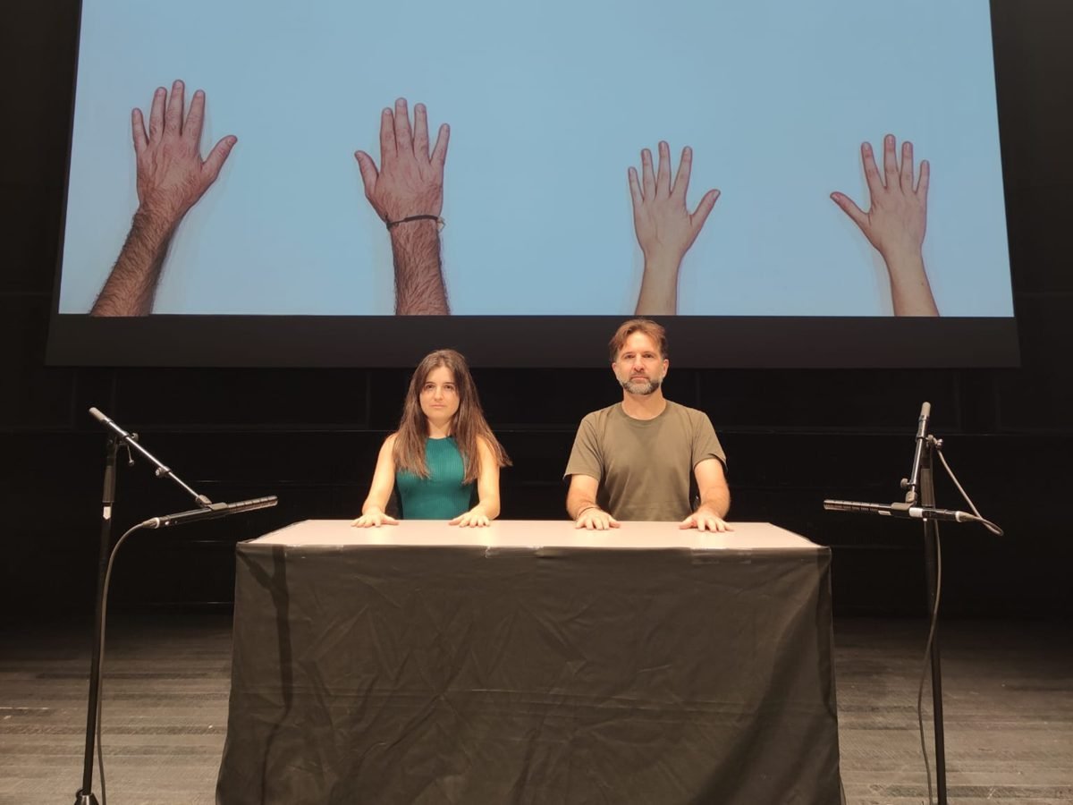 Gemma Galeano and Diego Kohn at the table; on the screen above, four open hands