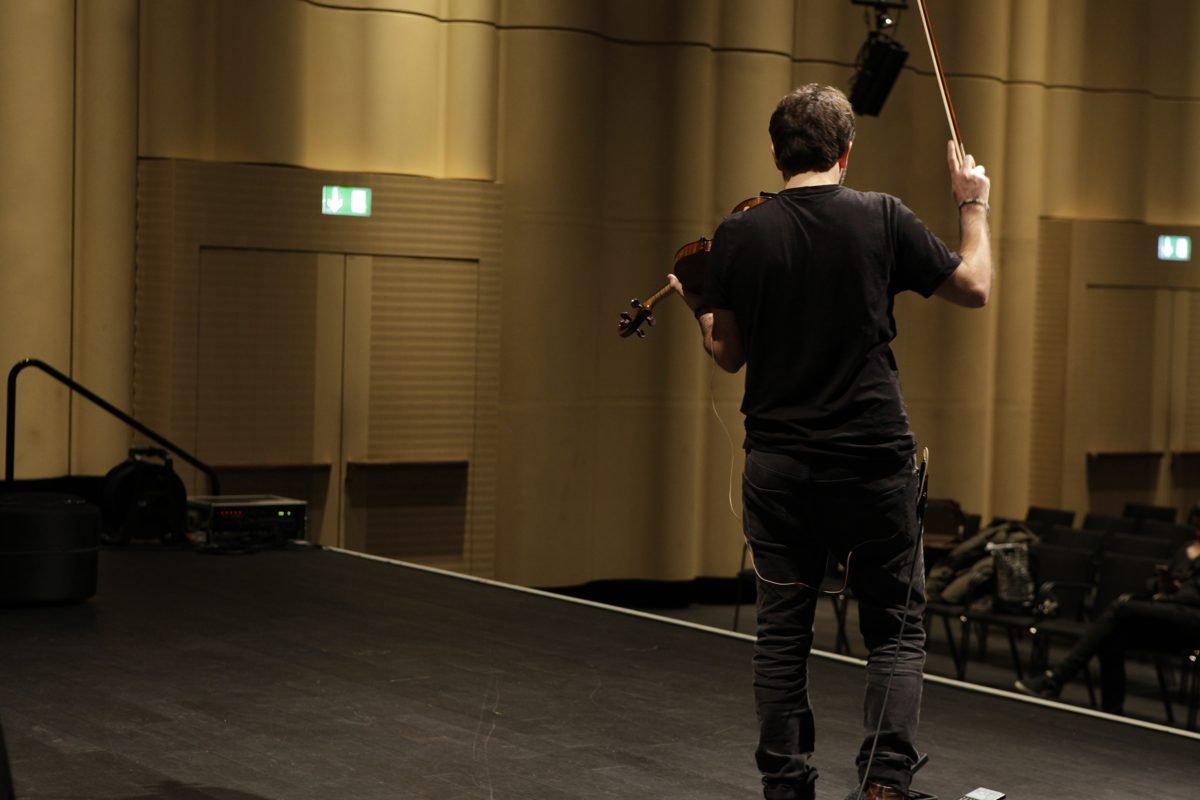 Diego Kohn from behind, bow raised, a loudspeaker hanging above