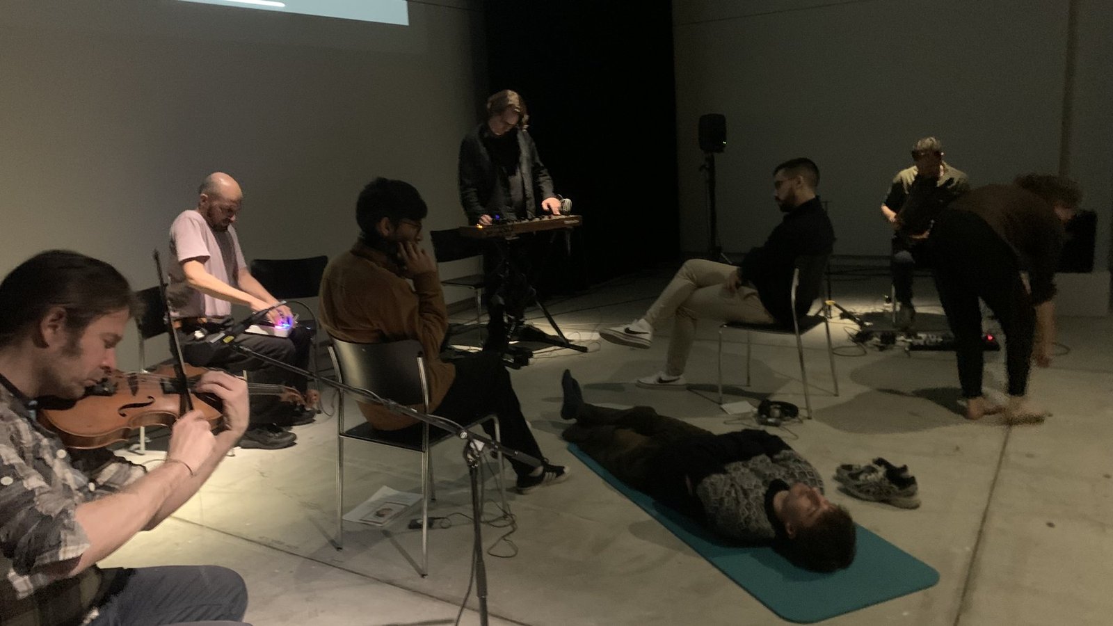 Listeners lying and sitting on the floor of a dim hall while musicians play among them