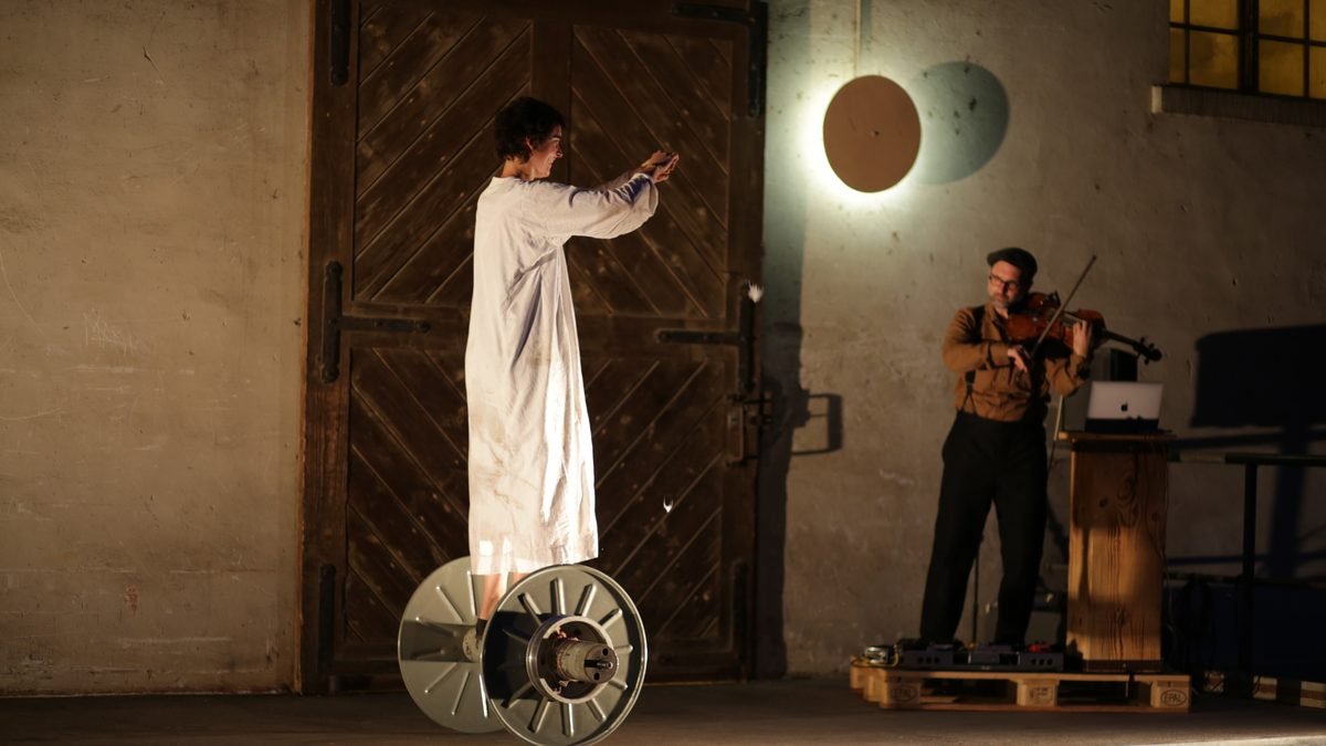 A dancer balancing on a large spool next to a viola player on a factory loading ramp