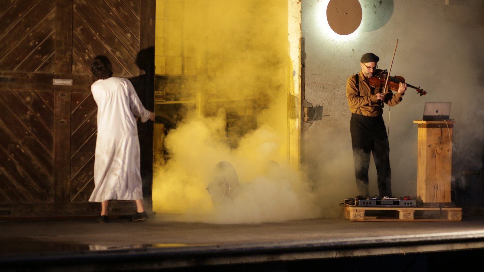 A dancer in a white work coat at an open factory door filled with yellow smoke; a viola player with a laptop and pedals plays beside her